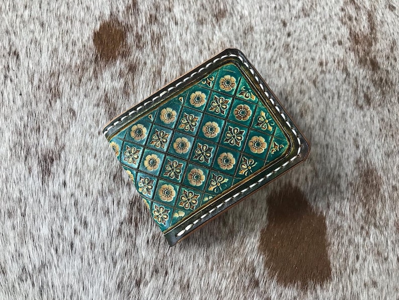 May include: A teal leather wallet with a gold and green geometric pattern. The wallet has a stitched edge and is closed.