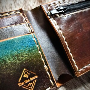 May include: A brown leather wallet with a zippered pocket and multiple card slots. The wallet has white stitching and a logo that reads "The Shoe Room".