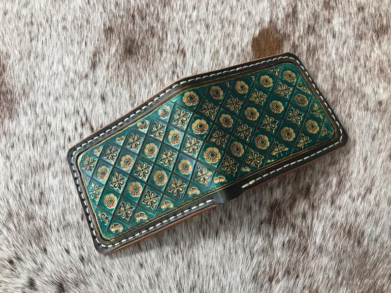 May include: A green leather wallet with a gold and green patterned design. The wallet has a white stitch around the edges.