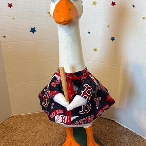 May include: A decorative goose figurine wearing a navy blue baseball cap with a red "B" logo and a black and red baseball jersey. The goose holds a wooden baseball bat and has orange feet and beak.