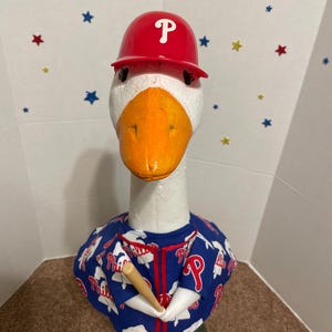 May include: A decorative goose figurine wearing a red baseball helmet with a white "P" and a blue jersey with "Phillies" text. The goose holds a wooden baseball bat. The background has star decorations.