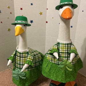 May include: Two goose figurines dressed in St. Patrick's Day attire. Each goose wears a green plaid dress, a green hat, and holds a shamrock. The geese have white bodies, orange beaks, and green feet.