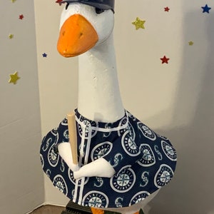 May include: A decorative white goose figurine wearing a navy blue baseball cap and a matching jersey with the Seattle Mariners logo. The goose holds a wooden baseball bat, with orange feet and beak.
