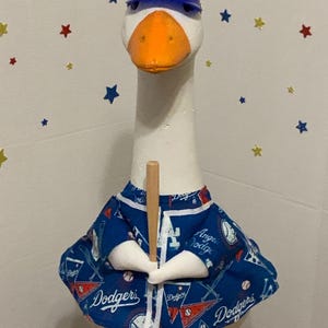 May include: A white goose figurine wearing a blue baseball cap with the letters "LA" and a blue Dodgers jersey. The goose is holding a wooden baseball bat. The background has star-shaped decorations.