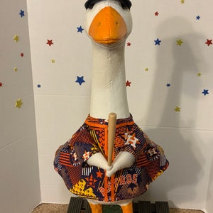 May include: A decorative goose figurine wearing a Houston Astros baseball uniform, including a navy blue helmet with the Astros logo, a jersey, and holding a wooden baseball bat. The goose has orange feet and beak.
