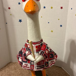 May include: A decorative goose figurine wearing a red baseball cap and a patterned shirt with the St. Louis Cardinals logo. The goose holds a wooden baseball bat, with orange feet and beak.