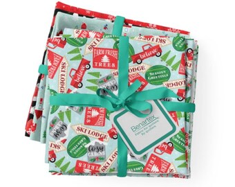 May include: A stack of four fabric squares with a Christmas theme. The fabric is blue with a white snowflake pattern and features red trucks, green trees, and signs with phrases like "Ski Lodge", "Cozy Cabins", and "Farm Fresh Trees". The fabric is tied with a green ribbon.