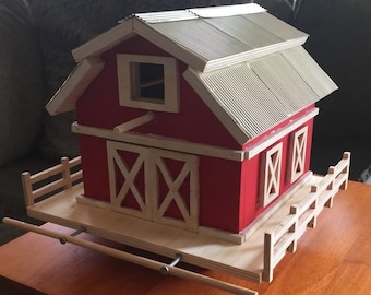 May include: A red wooden toy barn with a white roof and a white picket fence. The barn has a large double door with a white X pattern and two smaller windows with white X patterns. The barn is on a wooden base with a wooden rail.