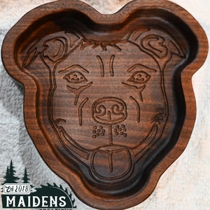 May include: A wooden bowl with a carved image of a dog's face. The bowl is dark brown and has a smooth, rounded shape. The dog's face is detailed and includes its eyes, nose, and mouth. The bowl is made by Maidens Creations, Est. 2018.