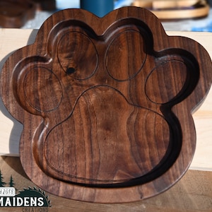 May include: A wooden paw print serving tray with four indentations. The tray is made of dark brown walnut wood and has a smooth, polished finish. The text "Est 2018 Maidens Creations" is engraved on the bottom of the tray.