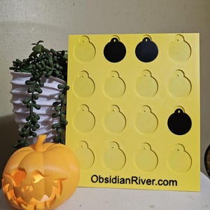 May include: A yellow pet tag display board with 16 tag-shaped slots, some filled with black tags. A carved orange pumpkin and a white planter with greenery are on the left. The website ObsidianRiver.com is printed on the board.