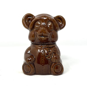 Puede incluir: Un tarro de miel de cerámica marrón en forma de oso con tapa cerrada. El oso lleva una pajarita marrón y está sentado sobre una superficie blanca.