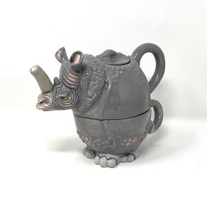 Può includere: Una teiera in ceramica grigia a forma di rinoceronte con una superficie testurizzata dettagliata. La teiera ha un beccuccio, un manico e un coperchio.