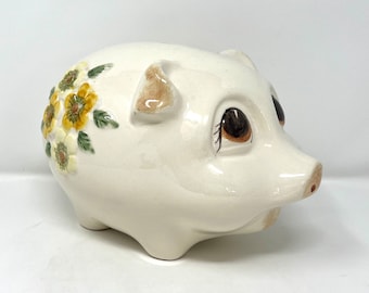 May include: White ceramic piggy bank with a floral design on the side. The flowers are yellow and green. The piggy bank has brown eyes and a small snout.