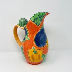 May include: A colourful ceramic jug with a handle, decorated with a variety of vegetables, including artichokes, peppers, and aubergines. The jug is painted in shades of orange, green, blue, and yellow.