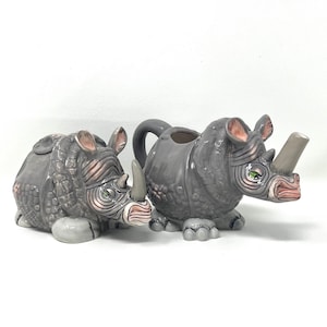 May include: A set of two ceramic rhinoceros teapots. The larger teapot has a spout and a handle. The smaller teapot is a creamer or sugar bowl. Both are gray with pink and green details.