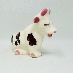 May include: A white ceramic cow figurine with brown spots and pink accents. The cow has closed eyes and a pink tongue sticking out.