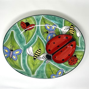 Könnte beinhalten: Eine grüne ovale Keramik-Servierplatte mit einem erhabenen Marienkäfer-Design. Der Marienkäfer ist rot mit schwarzen Punkten und hat schwarze Flügel. Die Platte ist mit grünen Blättern, Schmetterlingen und Bienen verziert.
