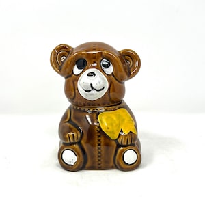 Puede incluir: Un tarro de miel de cerámica marrón en forma de oso de peluche con un parche amarillo en el pecho. El oso tiene ojos blancos, una nariz blanca y una boca blanca.