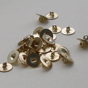 May include: A group of gold-coloured metal buttons with a loop on the top. The buttons are arranged in a pile on a white surface.