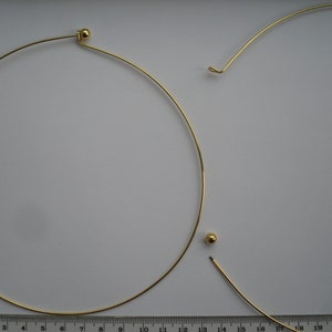 May include: Two gold-coloured metal choker necklaces with small round balls at the ends. The necklaces are on a white surface with a ruler for scale.