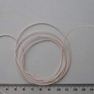 May include: A coil of pale pink thread, possibly for crafting or sewing, is shown against a light grey background. A ruler is positioned below the thread, providing a visual measurement reference. The thread is neatly coiled.