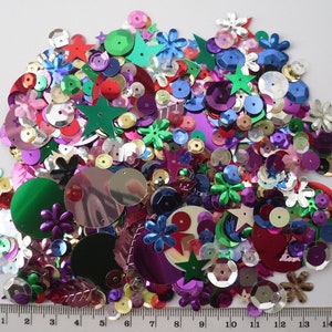 May include: A large assortment of colourful sequins in various shapes, including circles, stars, flowers, and leaves. The sequins are made of shiny, metallic material and come in a variety of colours, including red, green, blue, purple, gold, and silver.