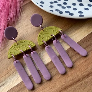 Può includere: Orecchini fatti a mano con un perno circolare viola. Gli orecchini hanno un semicerchio di argilla verde oliva con un motivo a spirale e tre pezzi rettangolari pendenti in una tonalità viola abbinata. Su una superficie di legno.