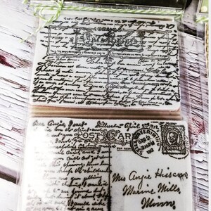 May include: Two vintage-style rubber stamps with black ink. The top stamp features a handwritten design with the text "Tous les pays derangery yeoptant pag la" and "Han forthen". The bottom stamp features a handwritten design with the text "Dear Angies Jones Millus fiel 2912" and "blindadAPTERIM". The stamp also features a vintage-style postage stamp with the text "POST CARD" and "FOR CORRESPONDENCE FOR ADDRESS".