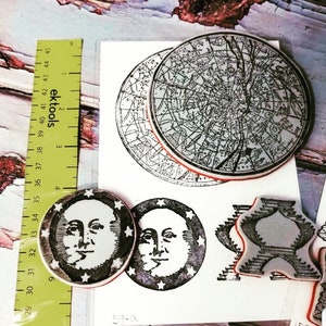 May include: Four rubber stamps with black ink on a white background. The stamps are of a celestial map, two moons with faces, and a star with a geometric design. The stamps are labeled with numbers 59-a and 159-6.