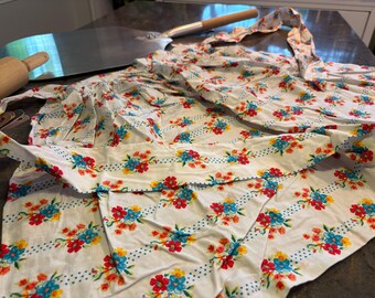 May include: White floral apron with red, blue, and yellow flowers. The apron has a ruffled edge and a dotted border. A wooden rolling pin and a metal pizza peel are in the background.