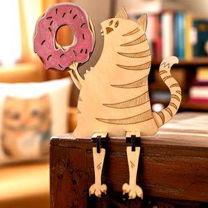 May include: A light-colored wooden cat figurine with dark etched stripes, holding a pink donut with dark sprinkles. The cat has dangling legs that hang over the edge of a surface, featuring a cutout design and a curved tail pointing upward.