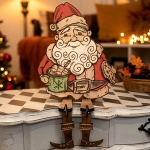 May include: A wooden cutout of Santa Claus wearing a red hat and coat, holding a green mug with a snowflake design and a cookie. The figure has a white beard, a belt, and dangling legs with brown boots, designed to sit on the edge of a surface.