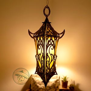 May include: A decorative hanging lantern with an intricate, dark brown wooden frame. The lantern has a pointed bottom and a loop at the top for hanging. The interior glows with a warm yellow light, creating a cozy ambiance. The design is reminiscent of a gothic style.