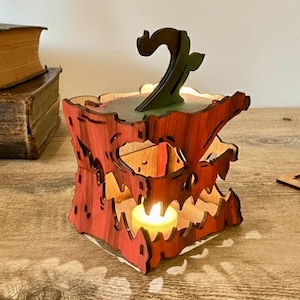 May include: A wooden, red-orange jack-o'-lantern candle holder with a green stem. The candle holder has a carved face and is lit from within by a small candle. The item is on a wooden surface, with other wooden cutouts and books in the background.
