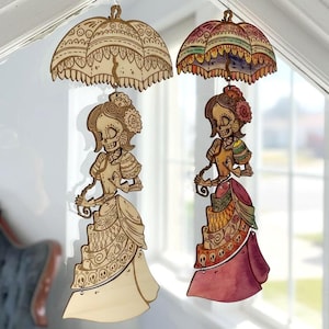 May include: Two hanging wooden ornaments featuring skeletal figures in Victorian-style dresses and umbrellas. One ornament is natural wood with laser-etched details, while the other is hand-painted in vibrant shades of pink, orange, and green.