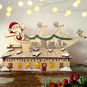Può includere: Una decorazione a forma di casa in legno con tetto chiaro e macchie di neve, una porta rossa e finestre illuminate. Sul tetto, un Babbo Natale in legno su una slitta è trainato da tre renne collegate da una ghirlanda color verde acqua.