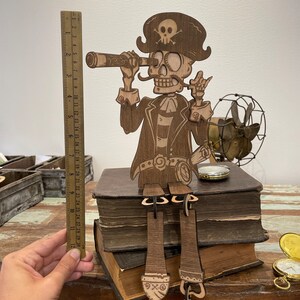May include: A laser-cut wooden pirate skeleton figure wearing a hat with a skull and crossbones, holding a telescope to one eye. The figure sits on two stacked books, with dangling legs. A ruler shows the figure is approximately 12 inches tall.