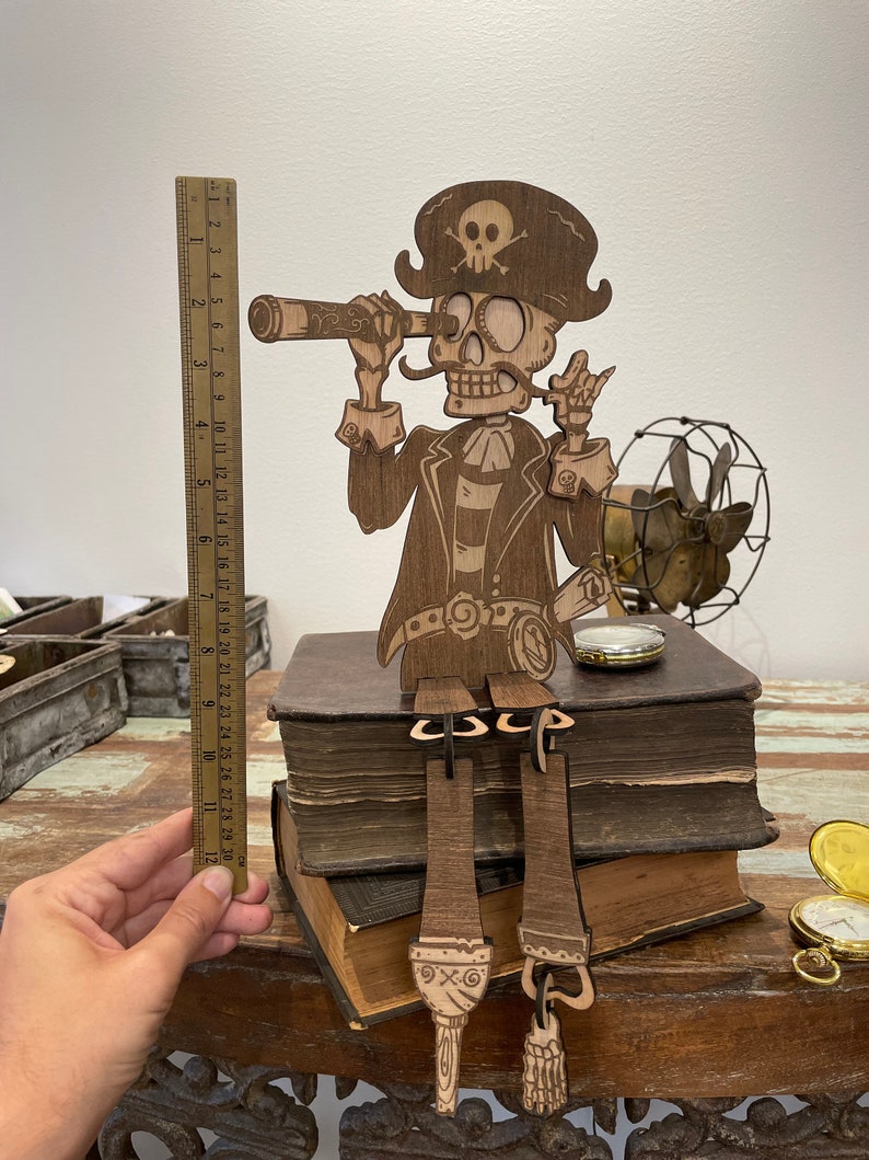 May include: A laser-cut wooden pirate skeleton figure wearing a hat with a skull and crossbones, holding a telescope to one eye. The figure sits on two stacked books, with dangling legs. A ruler shows the figure is approximately 12 inches tall.