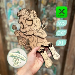 Pode incluir: Um recorte de madeira de um personagem de estilo de desenho animado costurado com uma cara de caveira e um chapéu. O personagem está segurando uma pequena figura costurada com uma cara de caveira. O recorte é segurado na mão de uma pessoa.