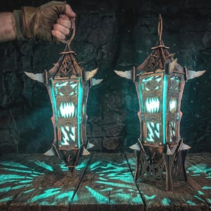 May include: Two dark brown, angular lanterns with pointed accents and glowing teal light emanating from cutouts featuring sharp, tooth-like shapes. One lantern is suspended by a ring at the top, while the other stands on a base.