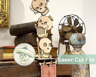May include: A vertical laser-cut wooden decoration featuring three stacked skulls, with the top skull holding maracas, the middle skull holding a taco, and the bottom skull holding a spear. A pink banner hangs below, and a circular inset shows a skull wearing a sombrero. The text Laser Cut File is visible.