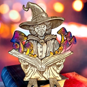 May include: Wooden witch figurine with a pointed hat, holding an open book with colorful mushrooms. The witch has a skull-shaped brooch. The mushrooms have purple, yellow, and blue caps. The figurine is on top of a stack of books.