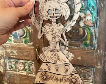 May include: A laser-cut wooden wall hanging featuring a skeletal figure with flowing hair, holding a rose. The figure is framed by a circular border and wears a multi-tiered dress decorated with engraved floral patterns and small skull motifs.