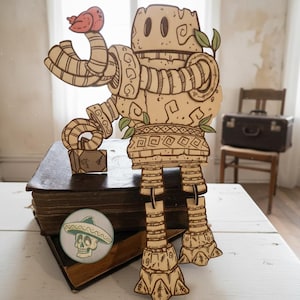 May include: A laser-cut wooden figure of a stone-like robot with segmented arms and legs, featuring a small red bird perched on its hand and a skull wearing a sombrero printed on a circular button placed on the books below the robot.