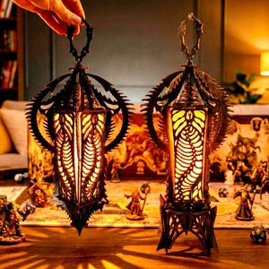 Puede incluir: Dos faroles ornamentados de metal oscuro con un interior cálido y brillante. Los faroles tienen un diseño detallado en forma de engranaje en la parte superior y los lados, con un asa en la parte superior. Los faroles están sobre una mesa con figuras en miniatura y un tablero de juego.