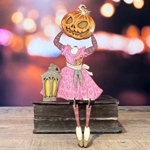 Könnte beinhalten: Eine dekorative Ausschnittfigur mit einem kopflosen Oberkörper in einem rosa Kleid mit Totenkopfmuster, die einen geschnitzten orangefarbenen Kürbis über dem Hals hält. Links steht eine gelbe Laterne und gestreifte Beine hängen herab.