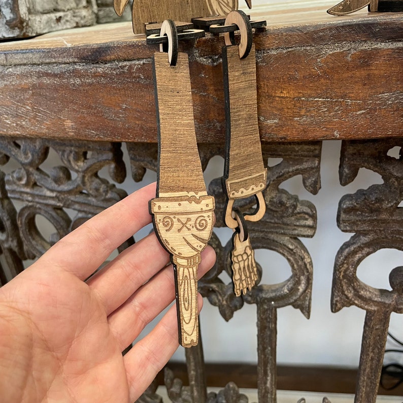 May include: A laser-cut wooden ornament featuring a dangling leg and foot design. The piece has a light brown wood grain finish with dark brown etched details, including a circular knee joint, a stylized lower leg, and a skeletal foot attachment.