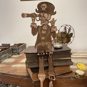 May include: A laser-cut wooden pirate skeleton figure wearing a hat with a skull and crossbones, holding a telescope to its eye. The figure sits on two stacked books, with dangling legs and feet. A metal ruler above shows a height of 11 inches.