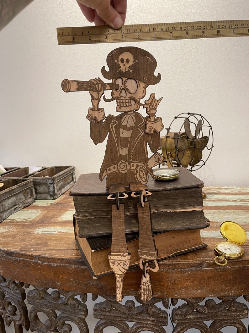 May include: A laser-cut wooden pirate skeleton figure wearing a hat with a skull and crossbones, holding a telescope to its eye. The figure sits on two stacked books, with dangling legs and feet. A metal ruler above shows a height of 11 inches.